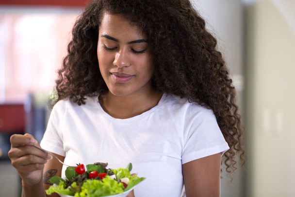 ホームキッチンで野菜サラダを食べる陽気な若いアフロアメリカ人女性 - Imagem. - 写真・画像
