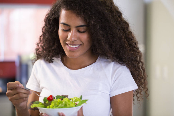 χαρούμενη νεαρή αφροαμερικάνικη γυναίκα που τρώει σαλάτα λαχανικών στο σπίτι κουζίνα-Απεικονίεμ. - Φωτογραφία, εικόνα