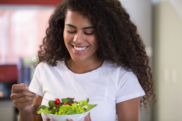 χαρούμενη νεαρή αφροαμερικάνικη γυναίκα που τρώει σαλάτα λαχανικών στο σπίτι κουζίνα-Απεικονίεμ. - Φωτογραφία, εικόνα
