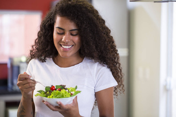 vrolijke jonge Afro-Amerikaanse vrouw die groente salade eet in huis keuken-Imagem. - Foto, afbeelding