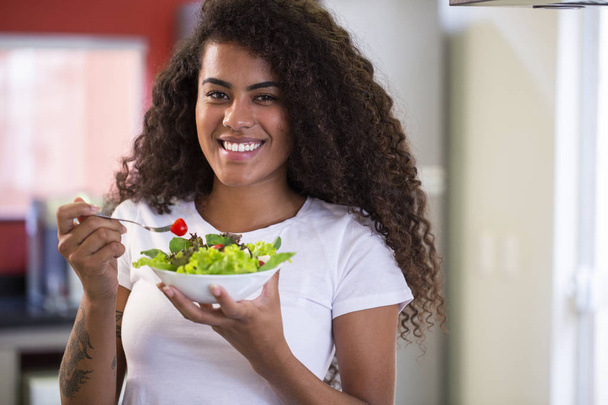 ホームキッチンで野菜サラダを食べる陽気な若いアフロアメリカ人女性 - Imagem. - 写真・画像