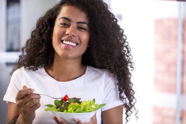 vrolijke jonge Afro-Amerikaanse vrouw die groente salade eet in huis keuken-Imagem. - Foto, afbeelding