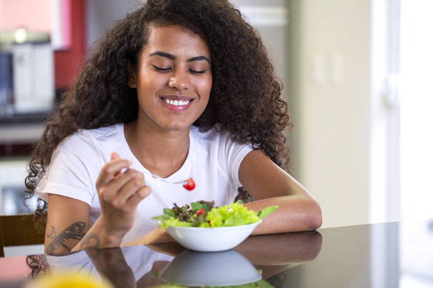 Wesoła młoda Afro amerykańska kobieta jedząca sałatkę warzywną w domowej kuchni-Imagem. - Zdjęcie, obraz