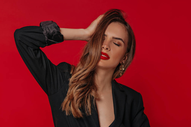 Glamorous lady with shiny hair and bright make up touching her hair and closed eyes with romantic smile over red background - Fotó, kép