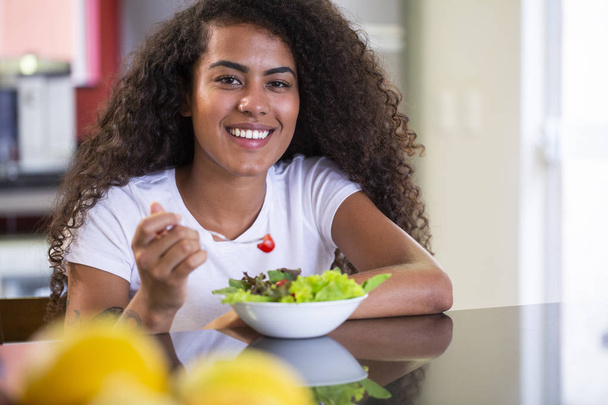 веселая молодая афро-американка ест овощной салат на домашней кухне
 - Фото, изображение