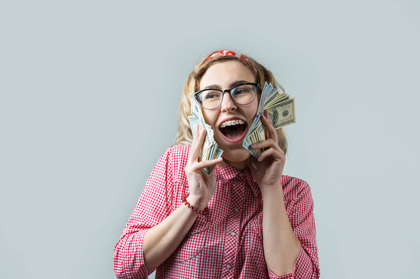 Κοντινά πλάνα της νεαρής όμορφη γυναίκα με ένα κόκκινο καρό πουκάμισο σε γυαλιά με αμερικανικά δολάρια χρήματα στο χέρι πάνω από γκρι φόντο. - Φωτογραφία, εικόνα