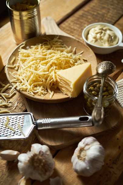 Shredded yellow cheese with cheese grater and capers and basil on kitchen table - Zdjęcie, obraz