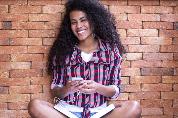 Happy student woman with afro hairstyle sitting with brick wall background using cell phone and smiling - Foto, immagini