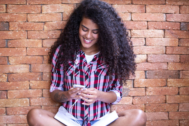 Happy student woman with afro hairstyle sitting with brick wall background using cell phone and smiling - Valokuva, kuva