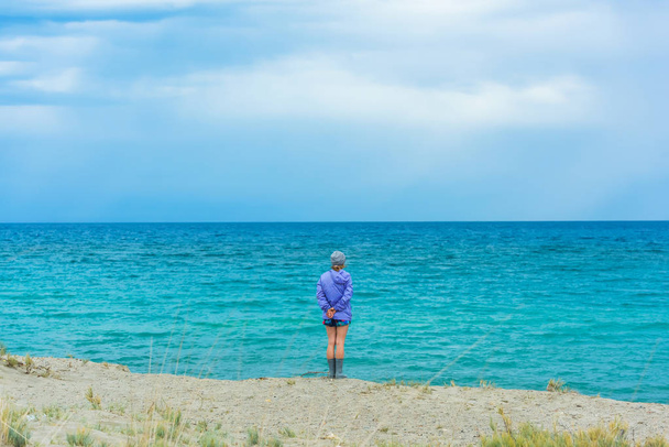 Een meisje in een paars jasje staat aan de oever van het meer Issyk-Kul. Rust in Kirgizië. Meisje op de achtergrond van een blauw meer. - Foto, afbeelding