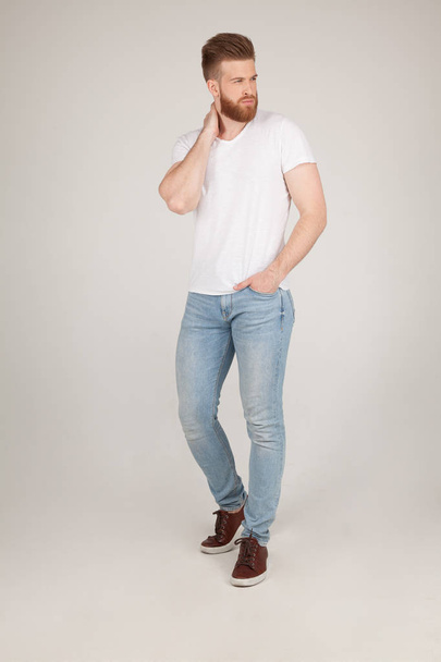 Homem barbudo sexy bonito com um estilo de cabelo bonito. posando na frente da câmera segurando uma mão em seu pescoço vestida com uma camiseta branca e jeans azul, posa na frente do fundo branco
. - Foto, Imagem