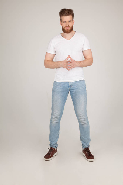 Beautiful sexy bearded man with a beautiful hair style. posing in front of the camera holding hands crossed in front of him. dressed in a white T shirt and blue jeans, poses in front of the white back - Photo, Image