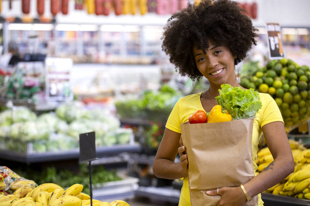 Healthy positive happy african woman holding a paper shopping bag full of fruit and vegetables. - Photo, Image