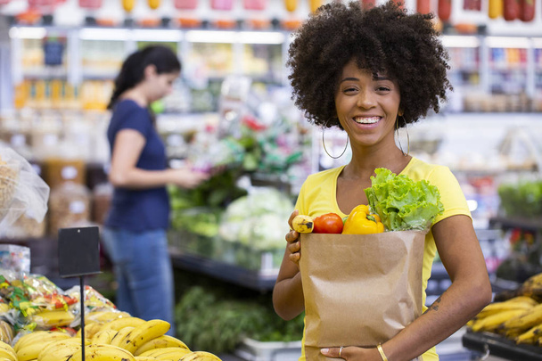 果物や野菜でいっぱいの紙の買い物袋を持つ健康的な肯定的な幸せなアフリカの女性. - 写真・画像