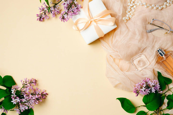 Feminine desk with silk scarf, gift box and lilac flowers on beige background. Top view, tender minimal flat lay style composition with copy space. - Foto, afbeelding