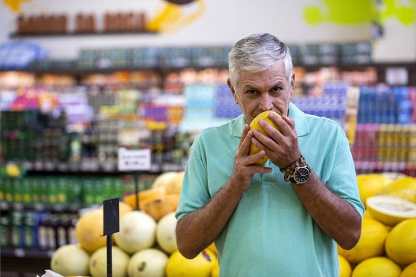 gutaussehender Mann, der in die Kamera blickt und eine Melone in der Hand hält. bärtige Kunden lächelnd. Abschnitt mit frischen Zitrusfrüchten im Hintergrund. - Foto, Bild