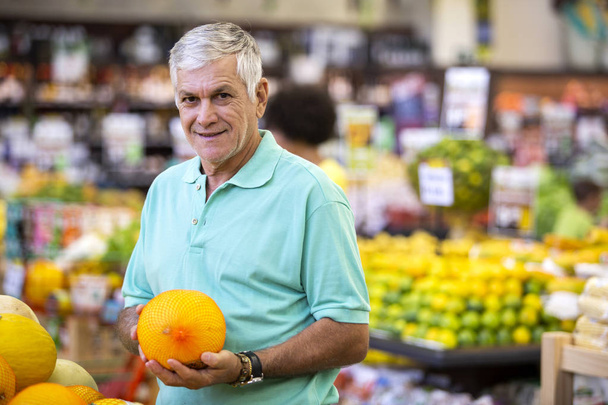 Handsome man posing, looking at camera and holding melon in hand. Bearded customer smiling. Section with fresh citruses on background. - Valokuva, kuva