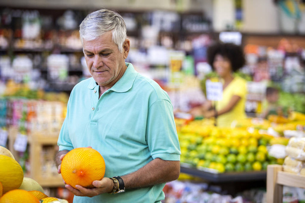 ハンサムな男は、果物を見て、手にメロンを保持し、ポーズ。あごひげを生やしたお客様の笑顔。背景に新鮮な柑橘類を持つセクション. - 写真・画像