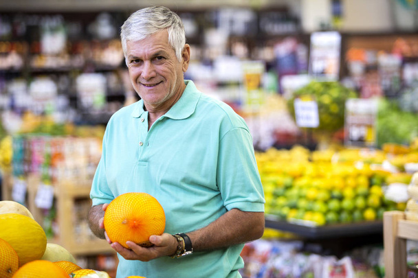 Handsome man posing, looking at camera and holding melon in hand. Bearded customer smiling. Section with fresh citruses on background. - Fotografie, Obrázek