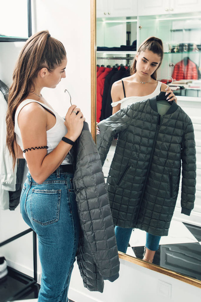 Young woman in a blu jeance white t-shirt trying on gray down coat in a luxury boutique - Photo, Image