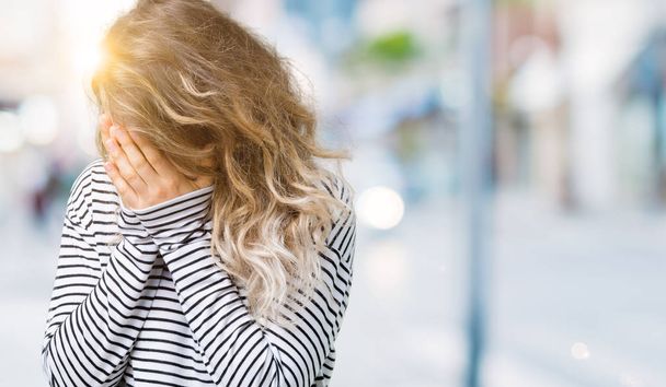 Krásná mladá blond žena na sobě pruhy svetr přes samostatný pozadí se smutným výrazem pokrývající obličej rukama a plakat. Koncept deprese. - Fotografie, Obrázek