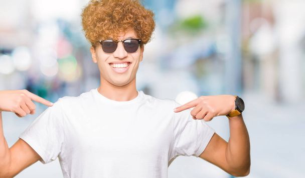 Jeune homme beau avec des cheveux afro portant des lunettes de soleil regardant confiant avec le sourire sur le visage, se montrant du doigt fier et heureux
. - Photo, image