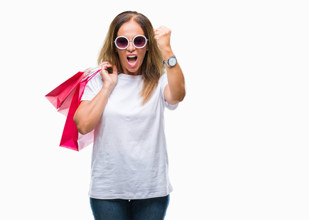 Mujer hispana de mediana edad sosteniendo bolsas de la compra en las ventas sobre fondo aislado molesto y frustrado gritando con ira, loco y gritando con la mano levantada, concepto de ira
 - Foto, Imagen