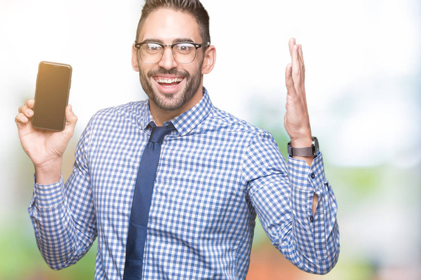 Young business man showing smartphone screen over isolated background very happy and excited, winner expression celebrating victory screaming with big smile and raised hands - Фото, зображення