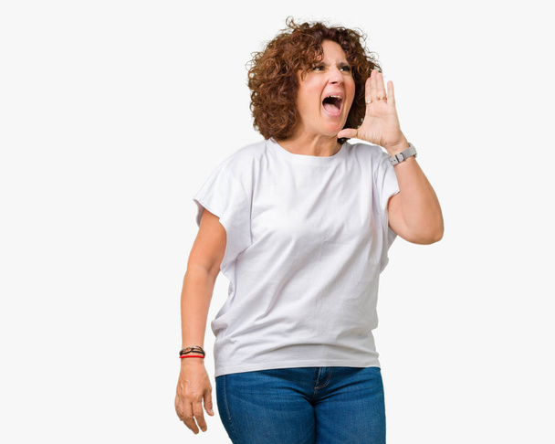 Piękny środkowy ager starszy kobieta nosi biały t-shirt na białym tle, krzycząc i krzycząc głośno na bok, z ręką na ustach. Komunikacja koncepcja. - Zdjęcie, obraz