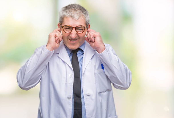 Bellissimo dottore anziano, scienziato professionista che indossa un camice bianco su sfondo isolato coprendo le orecchie con le dita con espressione infastidita per il rumore della musica ad alto volume. Concetto di sordo
. - Foto, immagini