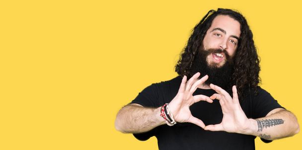 Young man with long hair and beard wearing heavy metal black outfit smiling in love showing heart symbol and shape with hands. Romantic concept. - Photo, Image