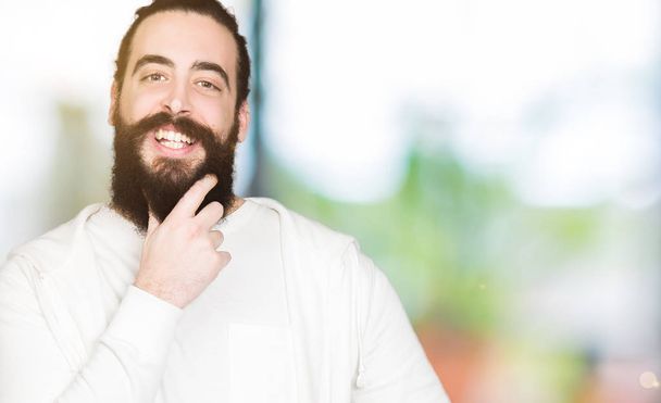 Young man with long hair and beard wearing sporty sweatshirt looking confident at the camera with smile with crossed arms and hand raised on chin. Thinking positive. - Photo, Image