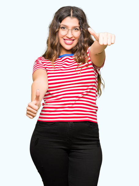 Young beautiful woman wearing glasses approving doing positive gesture with hand, thumbs up smiling and happy for success. Looking at the camera, winner gesture. - Foto, Bild