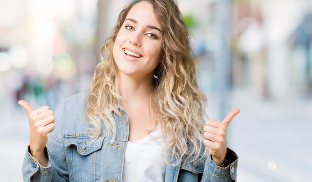 Beautiful young blonde woman wearing denim jacket over isolated background success sign doing positive gesture with hand, thumbs up smiling and happy. Looking at the camera with cheerful expression, winner gesture. - Photo, Image