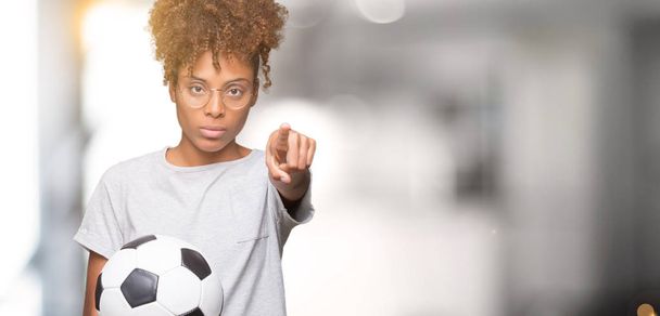 Молодая африканская американка держит футбольный мяч на изолированном фоне, указывая пальцем на камеру и на вас, знак руки, позитивный и уверенный жест спереди
 - Фото, изображение