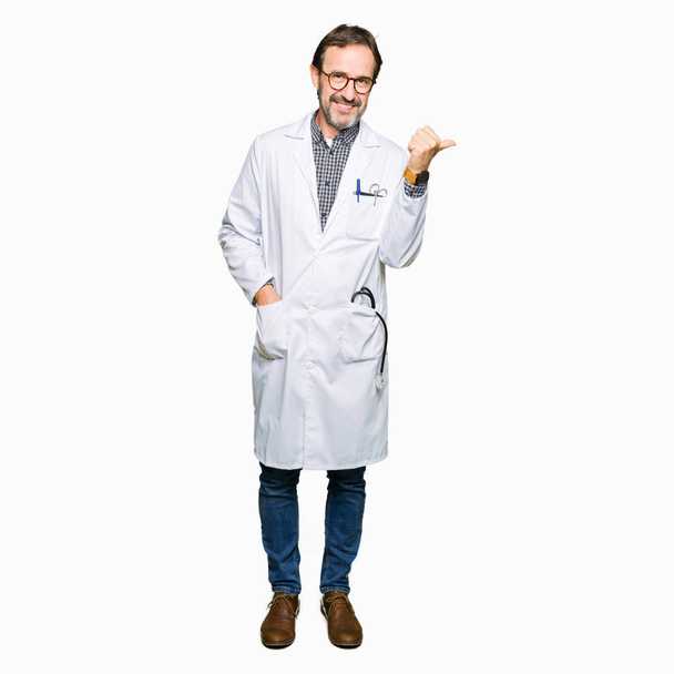 Középkorú orvos férfi visel orvosi kabátot mosolyogva boldog arcát keres, és rámutatva, hogy az oldalsó a hüvelykujj fel. - Fotó, kép