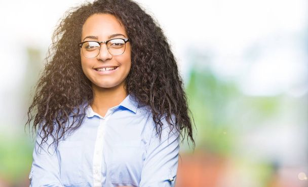 Young beautiful business girl with curly hair wearing glasses happy face smiling with crossed arms looking at the camera. Positive person. - 写真・画像