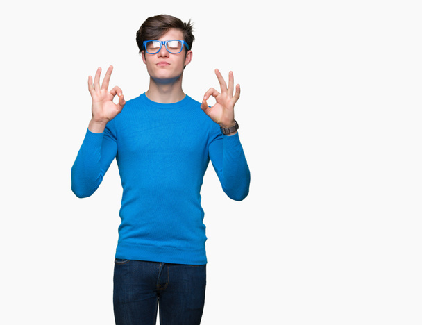 junger gutaussehender Mann mit blauer Brille über isoliertem Hintergrund entspannt und lächelt mit geschlossenen Augen und macht Meditationsgeste mit den Fingern. Yoga-Konzept. - Foto, Bild