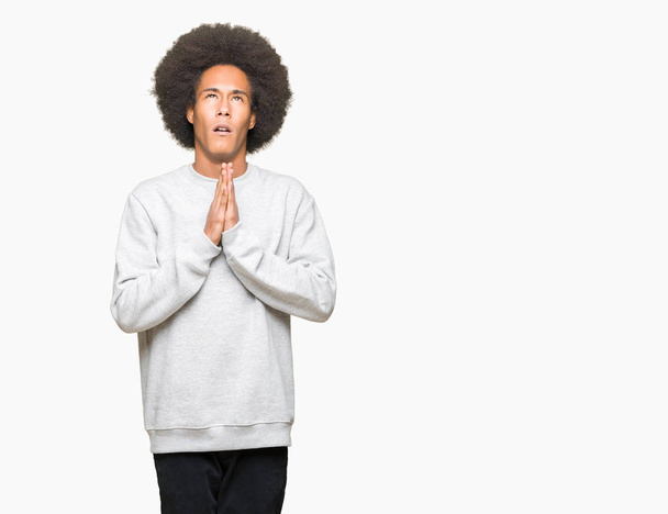 スポーティーなスウェットシャツを着たアフロヘアの若いアフリカ系アメリカ人男性は、非常に感情的で心配している顔に希望の表情と手をつないで祈ります。許しを請う宗教概念. - 写真・画像