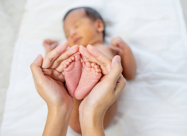 Mother hands hold little newborn baby feet with love emotion and the baby is sleeping on white bed - Φωτογραφία, εικόνα