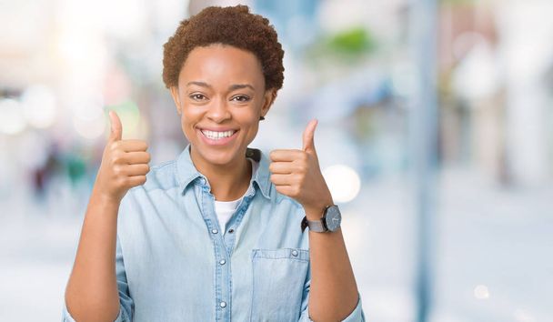 Young beautiful african american woman over isolated background success sign doing positive gesture with hand, thumbs up smiling and happy. Looking at the camera with cheerful expression, winner gesture. - Photo, Image