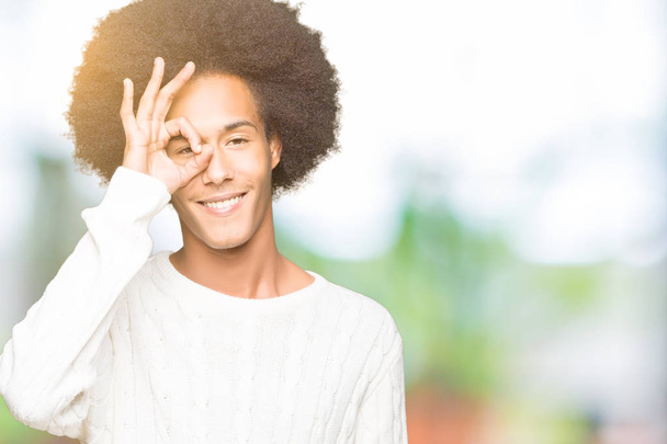 Jovem americano africano com cabelo afro vestindo camisola de inverno fazendo gesto ok com a mão sorrindo, olho olhando através dos dedos com rosto feliz
. - Foto, Imagem