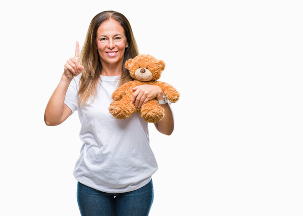 Middle age hispanic woman holding cute teddy bear plush over isolated background surprised with an idea or question pointing finger with happy face, number one - Photo, image