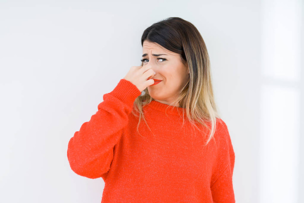 孤立した背景の上にカジュアルな赤いセーターを着ている若い女性は何か臭いと嫌な、耐え難い匂いを嗅ぎ、鼻の上に指で息を保持します。悪い匂いの概念. - 写真・画像