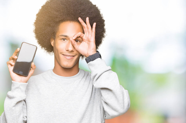 junger afrikanisch-amerikanischer Mann mit Afro-Haaren zeigt Smartphone-Bildschirm mit glücklichem Gesicht lächelnd tut ok Zeichen mit Hand auf Auge durch Finger schauen - Foto, Bild