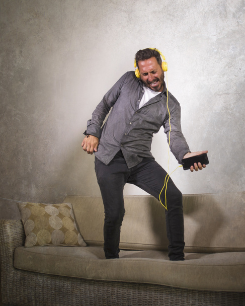 attraktiver Mann, der mit Kopfhörer und Handy Online-Musik hört, Luftgitarre spielt und unbeschwert auf dem Sofa im Wohnzimmer tanzt  - Foto, Bild