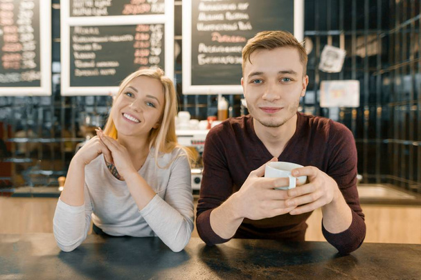 Young smiling couple of coffee shop workers, man and woman posing near the bar counter and coffee machine with cup of fresh coffee. Teamwork, staff, small business, people concept - Photo, image