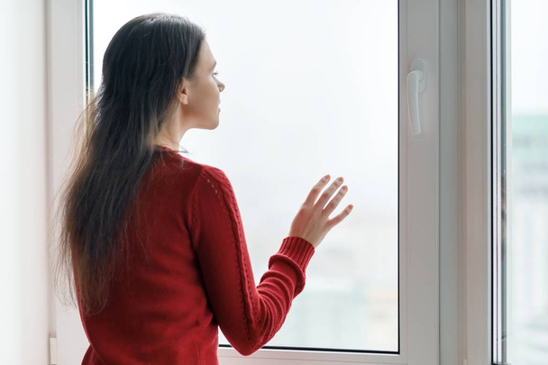 Young woman in red sweater looking out the window, female put her hands on the window glass, side view, window in skyscraper in morning on cloudy day - 写真・画像