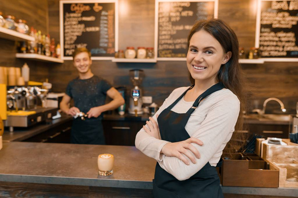 Portrait of smiling young female coffee shop owner, confident woman with arms crossed standing at the counter with barista working in background making drinks - Foto, immagini