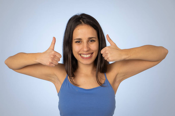 Portrait of beautiful young latin woman showing thumbs up sign feeling cheerful, happy and satisfied. Young happy student woman making thumb up in joyful approval gesture isolated on blue background. - Фото, изображение